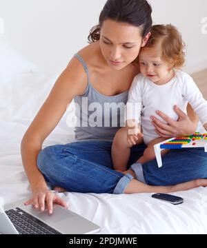 Aufwachsen in einer Welt der Technologie. Eine junge Mutter, die versucht, an ihrem Laptop zu arbeiten, während ihre kleine Tochter auf ihrem Schoß sitzt und sie beobachtet. Stockfoto