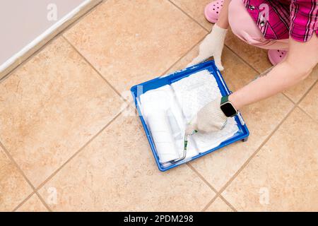 Woman-Wand- und Pinsel-Maler taucht Malerwalze mit weißer Farbe in eine Schale. Kosmetische Reparaturen in der Wohnung. Stockfoto