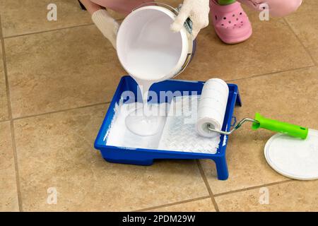 Die Wand- und Pinselmaler der Frau schüttet weiße Farbe in die Schale. Schließen. Kosmetische Reparaturen in der Wohnung. Stockfoto