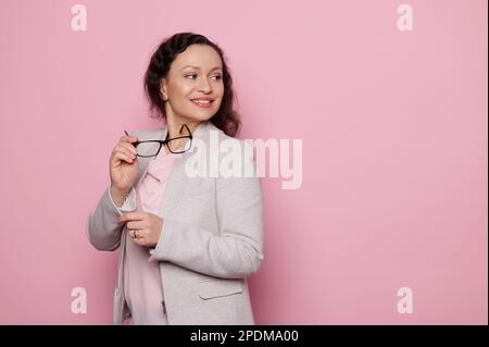 Selbstbewusste, schöne Frau in einem formellen Anzug, mit trendiger Brille, lächelnd auf den Kopierbereich auf pinkfarbenem Hintergrund Stockfoto