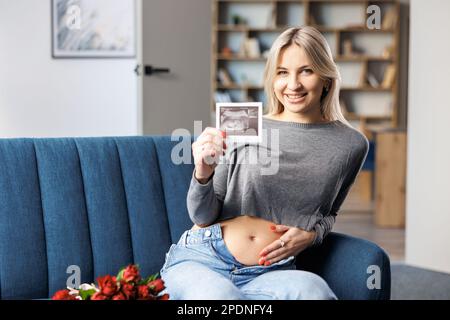 Foto einer schwangeren Frau zu Hause, die auf dem Sofa neben einem wunderschönen Blumenstrauß sitzt und in der Hand den Ultraschall-Scan-Druck des Babys hält Stockfoto