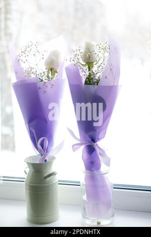 Zwei Sträuße weißer Rosen in lila Verpackung auf der Fensterbank Stockfoto