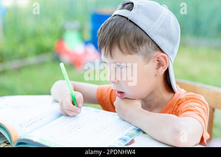 Ein Junge mit Mütze löst mathematische Beispiele an einem Tisch auf der Straße. Das Kind macht Hausaufgaben im Park Stockfoto