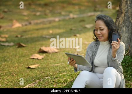 Positive Frau mittleren Alters, die an sonnigen, schönen Tagen im Park eine Papptasse Kaffee mit einem digitalen Tablet unter dem Baum hält Stockfoto