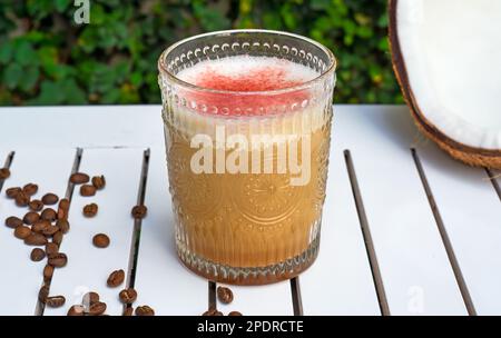 RAF-Kaffee Kokosnuss mit frischer Kokosnuss und Kaffeebohnen auf weißem Holztisch gegen grüne Blätter mit Kopierbereich Stockfoto