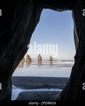 Blick auf die Three Sisters und den Berg Taranaki, eingerahmt von der Felshöhle am Strand von Tongaporutu. Taranaki. Vertikales Format. Stockfoto