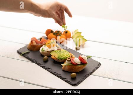 Zubereitung von Sandwiches, Zubereitung von Crostini, Canape, Antipasti mit Lachs Avocado, Tomaten, Frischkäse, Mozzarella für Party, Geburtstag. Das Konzept der gesunden Ernährung und der mediterranen Küche. Stockfoto