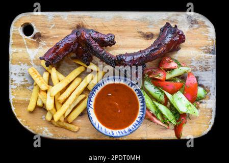 Gegrillte geräucherte Schweinerippchen mit pommes Frites, Sauce und Salat auf Holzbrett auf schwarzem Hintergrund, Draufsicht Stockfoto