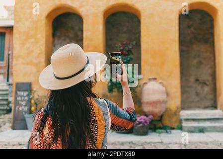 Eine Rückansicht einer Frau mit Sonnenhut, die ein Telefon hält und ein Foto des alten orangefarbenen Hauses im mediterranen Dorf macht Stockfoto