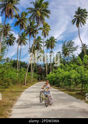 Vertikales Foto einer stilvollen Frau auf dem Fahrrad auf einem Sandweg, umgeben von hohen Palmen auf einer tropischen Insel Stockfoto
