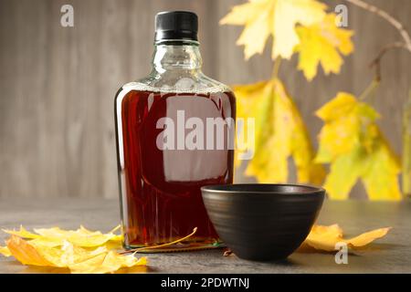 Flasche mit leckerem Ahornsirup, Schüssel und trockenen Blättern auf einem hellgrauen Tisch Stockfoto