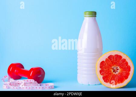 Minimalistisches Diät- und Sportkonzept. Rote Kurzhantel, Kefir-Flasche, Maßband auf blauem Hintergrund. Saftige Grapefruit für eine Sportdiät. Stockfoto