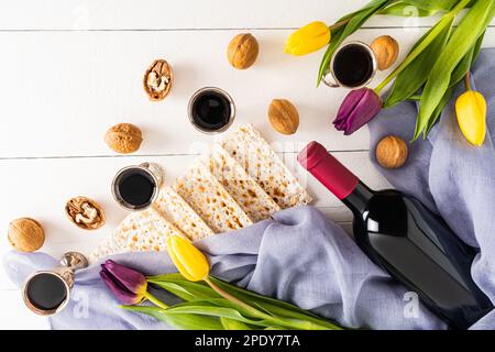 Silberne Gläser mit Rotwein, Matzah, eine Flasche Wein und Tulpen auf weißem Holzhintergrund. Das Konzept des jüdischen Passahurlaubs Stockfoto