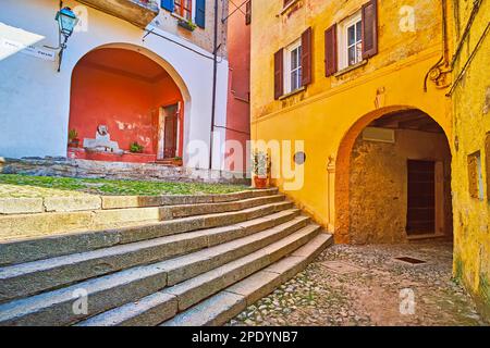 Der kleine Trinkbrunnen in Nischen, ein bogenförmiger Pass und schäbige Häuser auf der mittelalterlichen Piazza Paolo Pagani in Castello, Valsolda, Italien Stockfoto