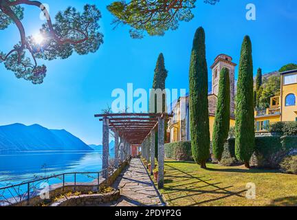 Der grüne Garten am Ufer des Luganer Sees mit wunderschönem Steinpergola-Fußweg und der mittelalterlichen Kirche San Sebastiano hinter den Zypressen, Oria, Vals Stockfoto