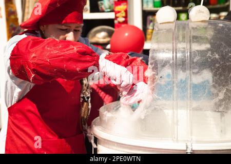 Khabarowsk, Russland - 1. Mai 2017: Mädchen in roter Uniform, die mit Zuckerwatte-Maschine arbeitet. Weibliche Lebensmittelverkäuferin, die Süßigkeiten-Zahnseide bei einer Fee verkauft. Straße f Stockfoto