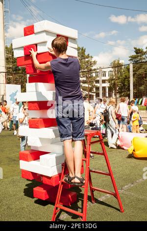 Chabarowsk, Russland - 13. August 2017: Junger Mann spielen Riesen jenga Spiel im Freien mit einer Leiter. Person ein Big Tower von roten und weißen Steinen pop Stockfoto