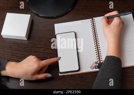 Weibliche Hände, ein Mädchen schreibt in einem Notebook, benutzt ein schwarzes Smartphone, Desktop, dunklen Hintergrund mit Kopierbereich, für Werbung, Draufsicht, weiße Box nic Stockfoto