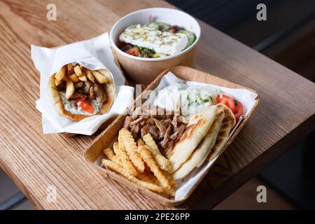 Griechisches Fast Food zum Mitnehmen. Traditionelles Pita Souvlaki, Gyros, griechischer Salat und Pita mit Pommes Frites, serviert in recyclingfähigem Papiergeschirr auf dem Tisch. Stockfoto