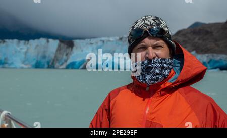 Junger Tourist mit roter Jacke, Schal und Hut, der an einem bewölkten Tag vorne auf einem Boot vor einem Gletscher im Hintergrund posiert Stockfoto