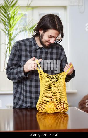 Junger Mann mit wiederverwendbarem Netzbeutel mit frischen Orangen und Äpfeln. Stockfoto