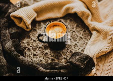 Unter den warmen Wollpulvern befindet sich eine schwarze Tasse mit heißem, starkem Kaffee. Trinken Sie in der kalten Jahreszeit. Komfort für Zuhause. Stockfoto