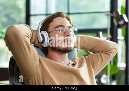 Nahaufnahme. Ein junger Freiberufler, ein Programmierer, sitzt im Büro und trägt weiße Kopfhörer, Hände hinter dem Kopf, Augen geschlossen. Ruht, misst, entspannt, hört Musik. Stockfoto