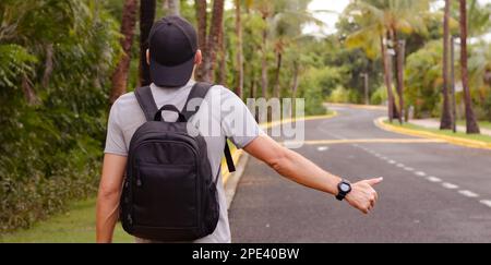 Anhalter mit Mütze und Rucksack, der auf der Straße steht und ein Auto fängt. Stockfoto