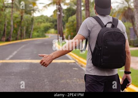 Hitchhiker auf einer leeren Straße in einem Land mit tropischem Klima. Ein Mann mit Blick nach hinten und Daumen-hoch-Anhalter. Stockfoto