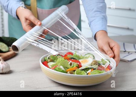 Eine Frau, die Plastikfolie über eine Schüssel frischen Salats auf einem Holztisch drinnen legt, Nahaufnahme Stockfoto