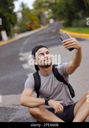 Junger Tourist, der auf der Straße sitzt und ein Selfie mit seinem Handy macht. Stockfoto