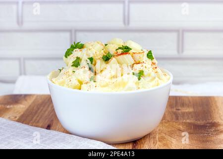 Frischer, hausgemachter traditioneller Amish-Kartoffelsalat mit Kartoffeln, gekochten Eiern, cremigem Dressing und Sellerie in einer weißen Schüssel auf Holzhintergrund Stockfoto