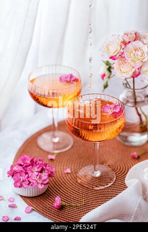 Tropfen fallen in den Cocktail, der mit Blumen auf einem weißen Tisch und einer Vase mit Rosen im Hintergrund dekoriert ist. Stockfoto