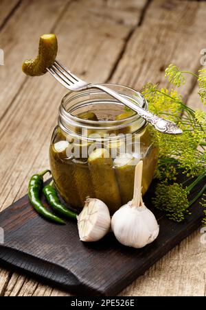 Eingelegte Gurken in einem transparenten Glasgefäß, eine Dosengurke auf einer alten Gabel, frischer Knoblauch, Kräuter und Gewürze auf Holzhintergrund. Stockfoto