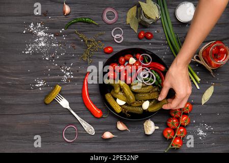 Ein Teller mit Gemüse in Dosen in den Händen auf dunklem Hintergrund. Eingelegte Kirschtomaten und Gewürzgurken in einem schwarzen Teller von oben. Eingelegtes Gemüse. Stockfoto