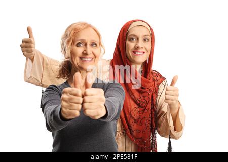 Glückliche reife Frau zeigt Daumen hoch und eine jüngere muslimische Frau, die sich hinter isoliert auf weißem Hintergrund posiert Stockfoto