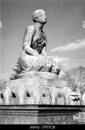 Eine verwitterte und abgenutzte Statue einer trauernden Frau auf dem Gipfel eines Grabdenkmals enthält ein Kreuz und Rosery, das dort von einem Besucher im Hintergrund von platziert wurde Stockfoto