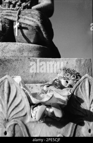Eine gruselige Puppe mit Dornenkrone und fehlenden Augen ruht auf einem gegebenen Denkmal mit einem Rosenkranz und Kreuz, das oben auf dem Woodbrook Friedhof Woburn hängt, Stockfoto