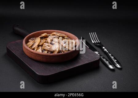 Gebratene oder gedünstete Champignon-Pilze in Scheiben mit Zwiebeln auf brauner Keramikplatte auf dunklem Betonhintergrund Stockfoto