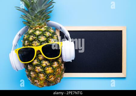 Ananas in Sonnenbrille und Kopfhörer mit leerer Tafel auf blauem Hintergrund Stockfoto