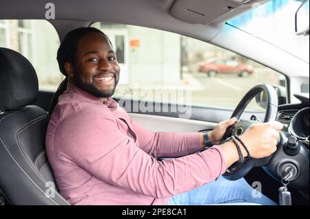 Ein glücklicher afroamerikanischer Mann mit den Dreadlocks fährt ein Auto, Seitenansicht von einem Beifahrersitz auf einen fröhlichen multirassischen Mann mit Lenkrad, kaufte ein Auto, lernte fahren Stockfoto