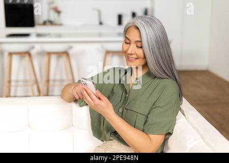 Lächelnde Frau mittleren Alters mit einem Smartphone sitzt auf dem Sofa zu Hause, ältere Dame hat angenehme Online-Chat auf einem Telefon, Scrollen News-Feed, verbringt Zeit in sozialen Netzwerken Stockfoto