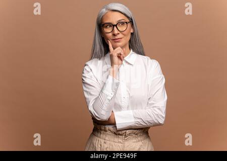 Porträt einer wohlüberlegten asiatischen Frau im weißen Hemd, die über ernste Probleme nachdenkt, mit unsicherem zögerlichen Ausdruck aussieht, eine schwierige Entscheidung trifft. Innenstudio isoliert auf beigefarbenem Hintergrund Stockfoto