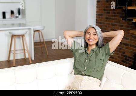 Eine Asiatin, die sich zu Hause ausruht und in die Kamera schaut. Positive Frau mittleren Alters mit langen Haaren auf dem Sofa in einem modernen Apartment. Das Konzept des Wohlbefindens Stockfoto