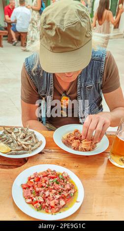Ein Mann, der in einem Fischrestaurant Fischgerichte mit Tintenfisch-Salat, kleinen gebratenen Fisch und gebratenen Tintenfisch zusammen mit Bier im Freien serviert Stockfoto