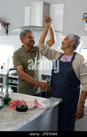 Ein birassistischer Senior-Mann tanzt mit einer Frau, die eine Schürze trägt, während er in der Küche steht, Kopierraum Stockfoto