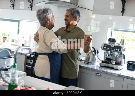 Glückliches, birassisches Seniorenpaar, das Händchen hält und sich ansieht, während er in der Küche tanzt Stockfoto