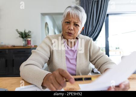 Birassistische Seniorin, die vor die Kamera schaut, Rechnungen auf dem Tisch analysiert und zu Hause sitzt Stockfoto