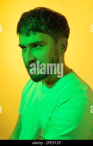 Kreatives Porträt. Lächelnder Mann mit einem schlauen Blick in grünen Neonlichtern auf gelbem Hintergrund. Moderner, gut aussehender Mann mittleren Alters mit Bart und Ohrring Stockfoto