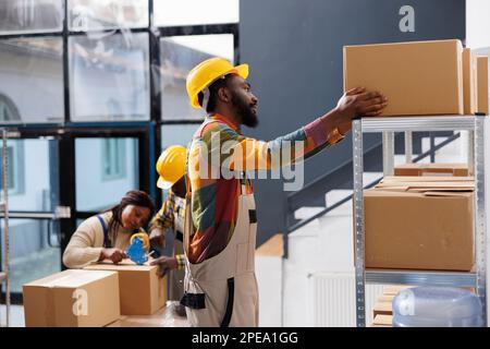 Lagerpacker wählt Pappkarton im Regal aus, während er auf der Leiter steht. Ein afroamerikanischer Lagerarbeiter bereitet das Kundenpaket vor und nimmt den Kartoncontainer mit Stockfoto